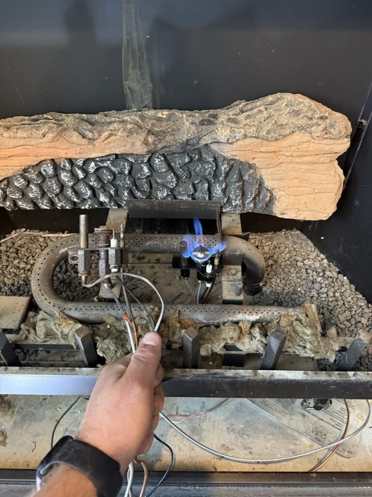 Removed fireplace pilot assembly held beside the installed pilot with a visible blue flame
